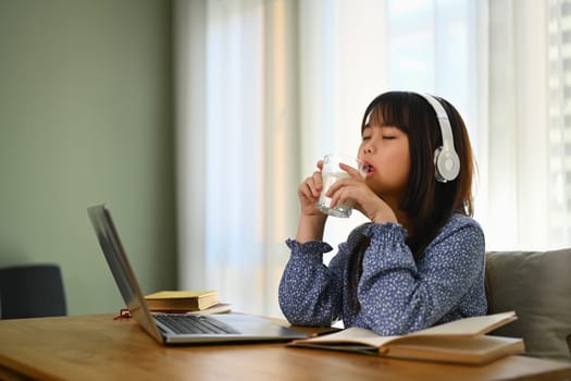 Asian schooler girl drinking milk while doing homework or studying online on laptop at home