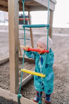 Little girl stands near a rope ladder holding on to a wooden crossbar. High quality photo