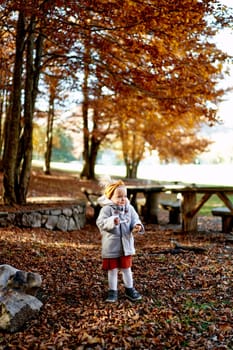 Little girl wraps herself in a coat while standing with a bun in her hand in an autumn park. High quality photo