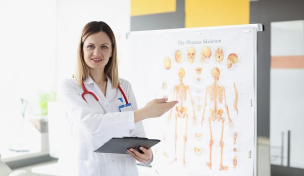 Doctor traumatologist holding medical documents and showing with his hand to poster with human skeleton. Medical education anatomy concept