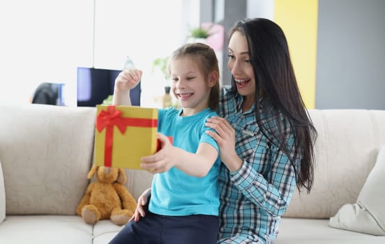 Little happy girl sitting on her mother lap and opening gift box at home. Selection of gifts for children concept