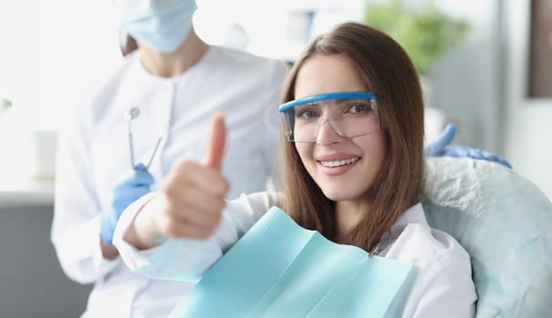Young woman in safety glasses sitting in dental chair and showing thumbs up. Quality dental care concept