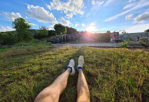 Male feet in white sneakers lying on green grass against background of waterfall closeup. Camping concept