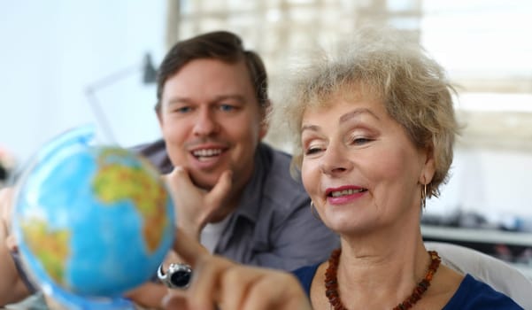Close-up of beautiful senior woman and middle-aged man explore blue globe. Mother and son choose country for future travel. Modern technology and leisure together concept