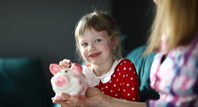 Portrait of smiling charming child holding piggy bank sitting on sofa indoors. Lovely blonde kid saving up money for future. Girl in red dress. Childhood and happiness concept