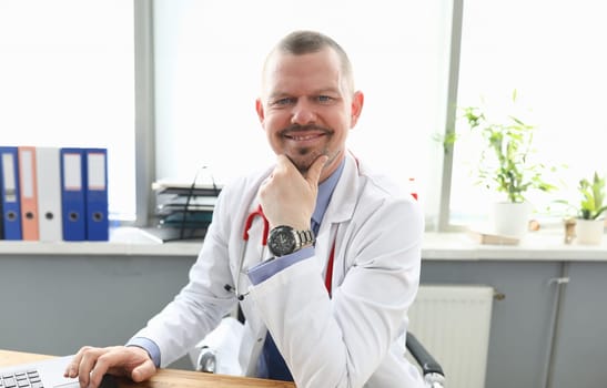 Portrait of happy cheerful professional doctor posing in office. Qualified male wearing uniform and red stethoscope. Laptop on wooden table. Medicine and healthcare concept
