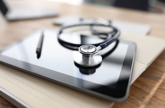 Close-up of modern black tablet device and doctors stethoscope. Silver pen on gadget. Macro shot medical equipment on desktop. Healthcare. Technology and medicine concept