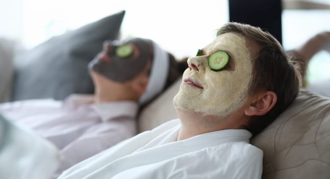 Close-up of wife and husband wearing cleaning and moisturizing face masks. Relaxed female and male laying on couch. Couple with cucumber slices on eyes. Leisure and cosmetology concept