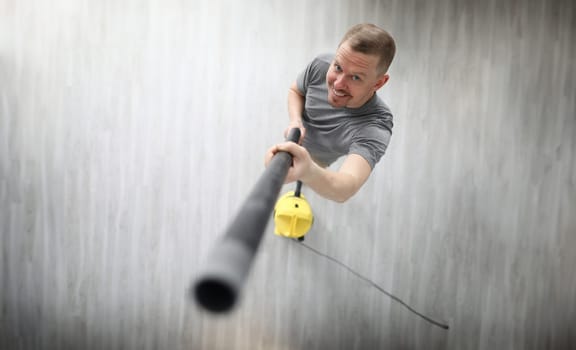 Top view of smiling middle-aged man cleaning apartment with grey vacuum cleaner. Cheerful person in grey shirt indoors. Renovation and construction site concept