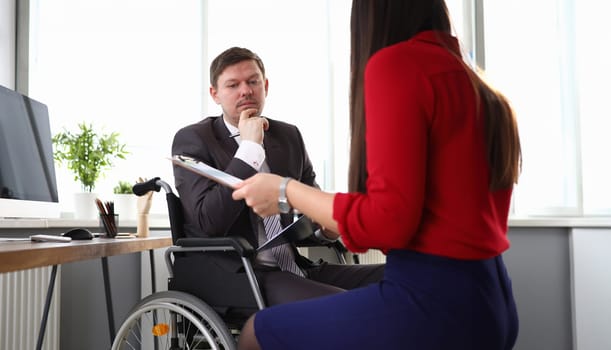 Portrait of male person sitting in wheelchair. Woman in red blouse holding clipboard for sign. Modern office with technology. Career help. Business and disabled people concept