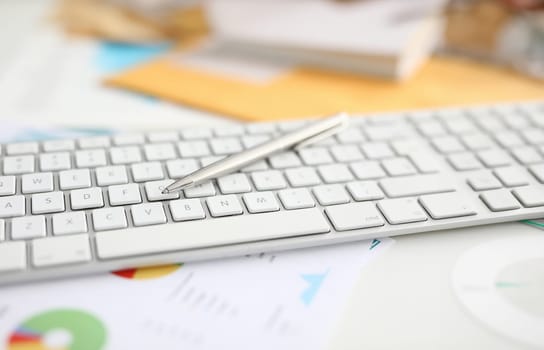 Close-up of white modern laptop keyboard on table. Silver pen for writing. Paper with chart in office. Executive manager desktop. Business cabinet and personal stuff concept