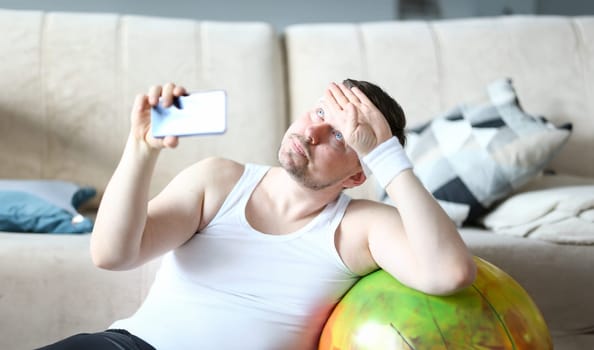 Thoughtful Man Shooting Training on Mobile Phone. Tired Sportsman Recording Video on Cellphone. Bearded and Overweight Male Using Smartphone to Calculate Calories. Guy Taking Photo on Fitness Ball