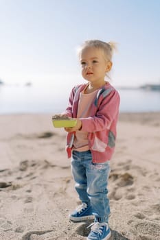 Little girl stands on the seashore with a shovel of sand in her hands. High quality photo
