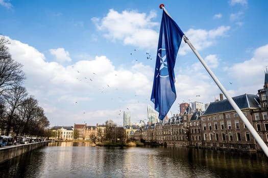 NATO flag in the center of Hague sky with dutch river channels and old city buildings view. Holland sityscape,