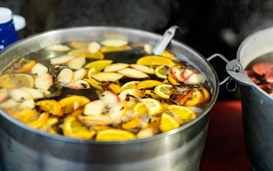 Beverage apples and oranges hot mulled wine in pot in street food market. Colorful hot fruit and berries drink at fair festival outdoors closeup