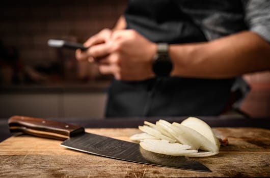 Chief hands holding smartphone at kitchen and sliced onion on wooden board. Man during cooking break with mobile cell phone and bitter vegetable