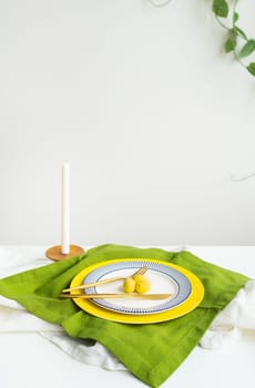 Minimalist table setting with green napkin, round plate with blue and yellow border, pair of fork and knife, white candle in background. Decor for the Easter table