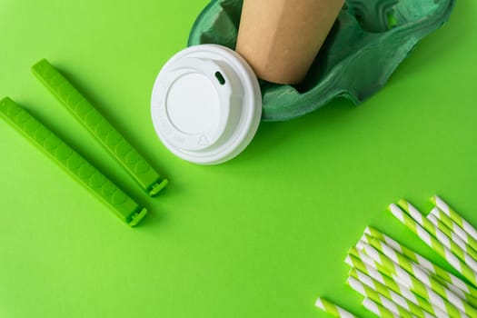 This image features an eco-friendly disposable coffee cup with green silicone straps and paper straws against a vibrant green background. The products are made from biodegradable materials