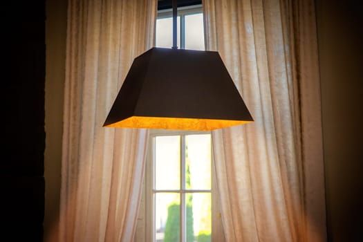 A lamp hanging from the ceiling shines with warm light in front of a window with curtains.