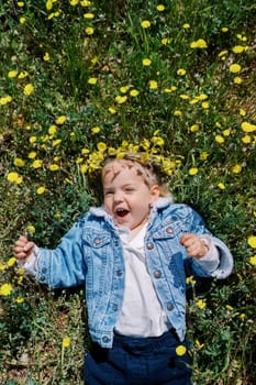Laughing little girl in a wreath with a flower in her hand lies on a flowering lawn. Top view. High quality photo
