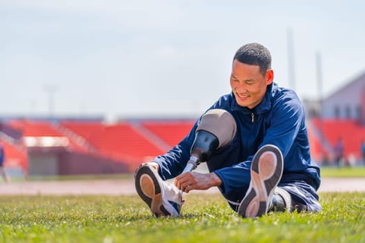 Sport man athlete with a prosthesis on his leg sit on grass field to tie shoelaces near the track at the stadium. Sport concept.
