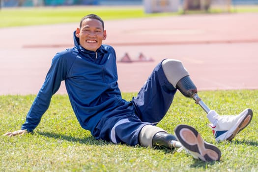 Close up sport man athlete with a prosthesis on his leg sit on grass field near track and look at camera with smiling to relax after exercise at the stadium. Sport concept.