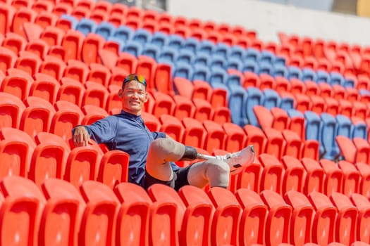 Sport man athlete prosthesis legs is sitting cross-legged on chair of amphitheater also look at camera with smiling in the stadium with day light.