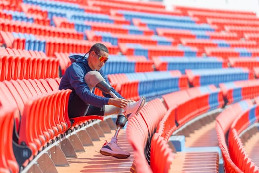 Sport man athlete with a prosthesis on his leg sit on chair of amphitheater to tie shoelaces in the stadium with day light.