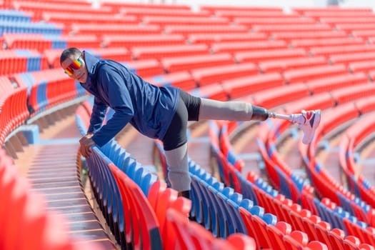Sport man athlete prosthesis legs use chair of amphitheater to warm up before exercise by raise his leg up and point to the back in the stadium with day light.
