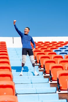 Vertical image of sport man athlete prosthesis legs stand on stairs with raise his hand up on amphitheater in the stadium with day light.