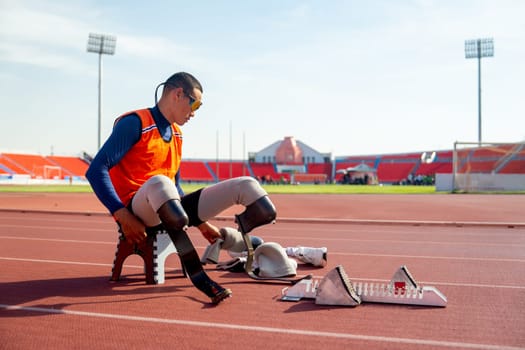 Sport man athlete with a prosthesis on his leg sit on small chair and prepare equipment for running at the stadium. Sport concept.