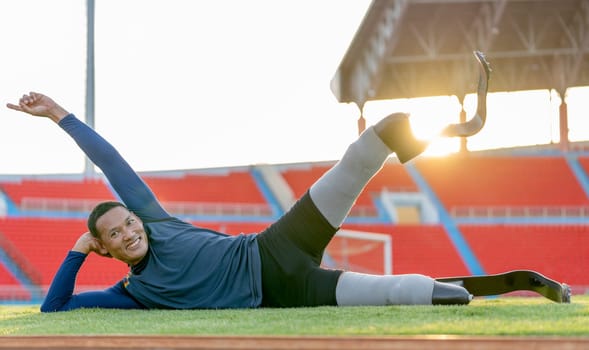 Sport man athlete prosthesis legs lie on his side down on grass field and raise one of his legs up also look at camera in the stadium with soft light in evening.