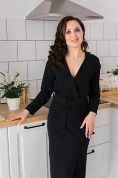 Beauty brunette woman standing in the kitchen. Attractive young girl smiling and touching the table. The girl looks to the side, elegant black and white dress