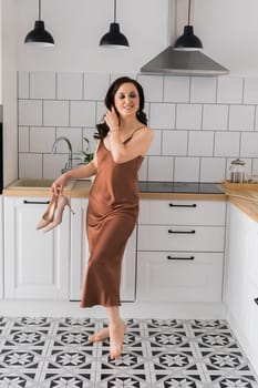 Smiling attractive young woman wearing silk long dress dancing at the kitchen at home