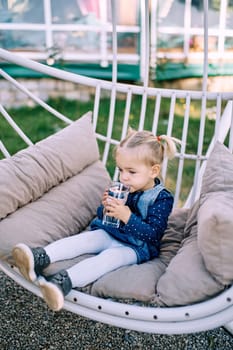 Little girl drinks water from a glass in her hands while sitting on a soft bench in the garden. High quality photo