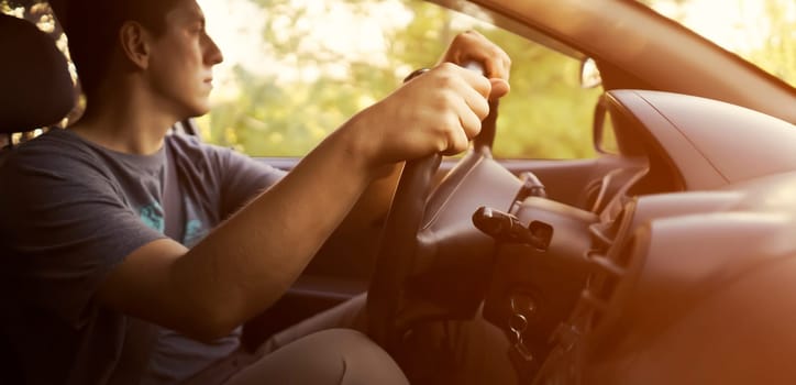 A young man travels in his car, drives along the road at sunset, holds the steering wheel with his hands, closeup view.