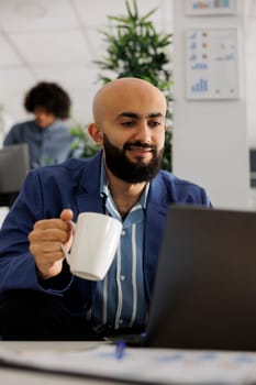 Smiling business company employee checking project financial report and drinking coffee. Arab start up entrepreneur holding tea mug and working at laptop in coworking space