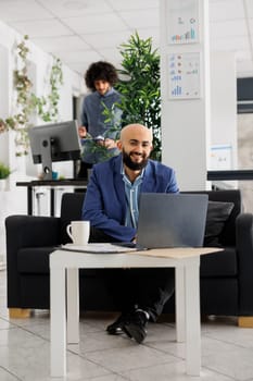 Smiling successful arab businessman working on laptop in coworking space portrait. Happy confident unicorn business entrepreneur sitting on couch at office workplace and looking at camera