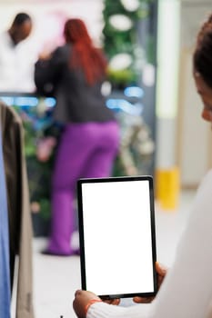 Woman using digital tablet mock up to browse clothing options in shopping mall. Boutique african american buyer holding portable gadget with blank white screen in vertical mode