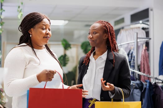 African american friends carrying shopping bags and chatting while browsing clothes in mall. Girlfriends holding paper packages while discussing fashion trends in boutique