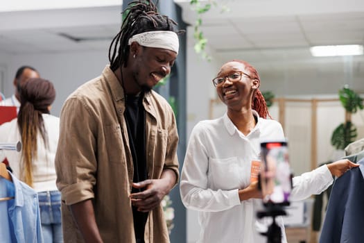 African american influencer and boutique owner live streaming on social media while showcasing outfits in showroom. Blogger and shop worker promoting fashion brand while recording video on smartphone