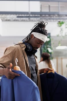 Customer holding two blazers and comparing style in fashion boutique. African american man examining trendy formal jackets on hangers before making purchase in shopping center