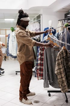 African american man browsing clothes in retail store showroom. Young customer checking apparel hanging on rack to buy casual plaid shirt in shopping mall fashion boutique