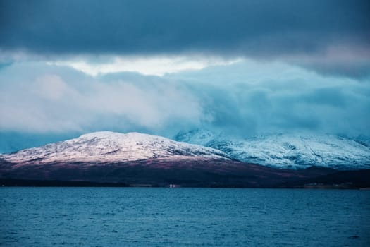 Scenery of snow-covered mountains across calm water, fjord shore near Tromso, Norway