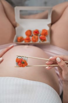 A pregnant woman sits on the sofa and eats rolls from a box. Food delivery. Top view of the tummy