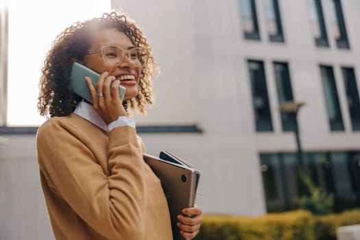 Pretty female entrepreneur is talking phone while standing with laptop on modern building background