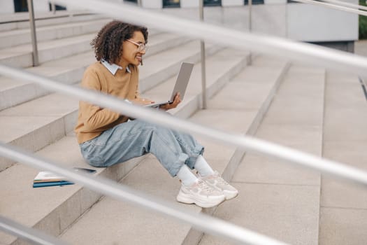 Smart woman student in eyeglasses is using pc laptop sitting outdoors on building background