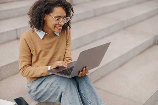 Smart woman student in eyeglasses is using pc laptop sitting outdoors on building background