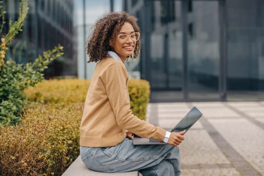 Cheerful woman manager in eyeglasses is working on laptop sitting on background of office building