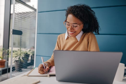 Smiling female analyst in eyeglasses working on laptop and making notes while sitting in cozy office